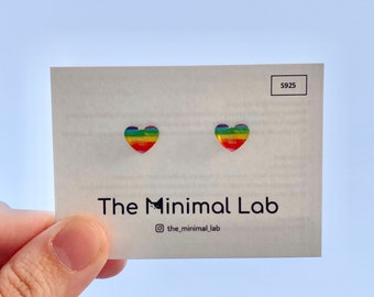 Puede incluir: Un par de pendientes de botón en forma de corazón arcoíris sobre una tarjeta blanca con el texto "The Minimal Lab" y "@the_minimal_lab". La tarjeta también tiene el número "S925" impreso en ella.