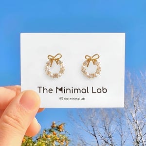 May include: A pair of gold-toned stud earrings shaped like wreaths, featuring small white pearls and clear crystals, topped with a gold bow. The earrings are displayed on a white card labeled The Minimal Lab with an Instagram handle.
