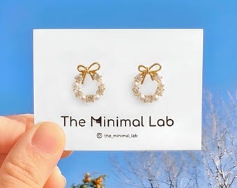 May include: A pair of gold-toned stud earrings shaped like wreaths, featuring small white pearls and clear crystals, topped with a gold bow. The earrings are displayed on a white card labeled The Minimal Lab with an Instagram handle.