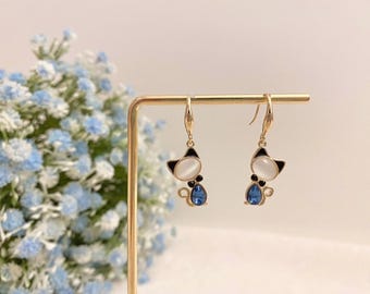 Puede incluir: Un par de pendientes colgantes de tono dorado con diseño de gato. Cada pendiente tiene una piedra cabujón ovalada blanca para la cabeza, orejas de esmalte negro, un pequeño acento negro y un cristal azul en forma de lágrima para el cuerpo.
