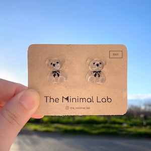 May include: A pair of clear resin teddy bear stud earrings with black bows. The earrings are displayed on a brown card with the text "The Minimal Lab" and "S925".