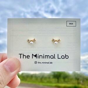 Puede incluir: Un par de pendientes de oro con una peque&ntilde;a perla y un dise&ntilde;o de lazo. Los pendientes est&aacute;n en una tarjeta blanca con el texto "The Minimal Lab" y "@the_minimal_lab".