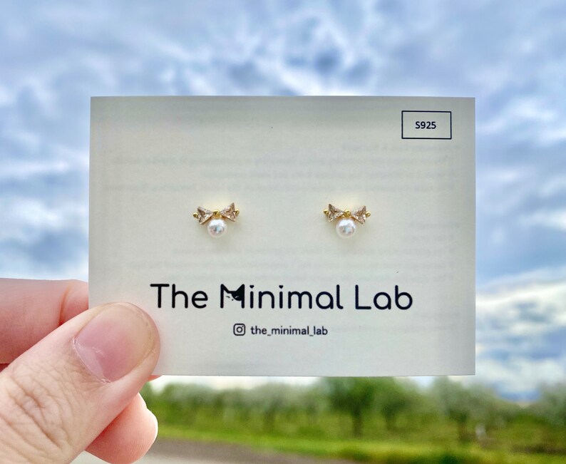 Puede incluir: Un par de pendientes de oro con una peque&ntilde;a perla y un dise&ntilde;o de lazo. Los pendientes est&aacute;n en una tarjeta blanca con el texto "The Minimal Lab" y "@the_minimal_lab".