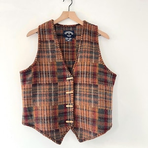 May include: A plaid vest in shades of brown, orange, and burgundy hangs on a wooden hanger. The vest has a V-neck and fastens with four wooden toggles. The fabric has a textured appearance.