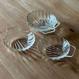 May include: Three clear glass bowls shaped like seashells with a ribbed texture. The bowls have handles on the sides.
