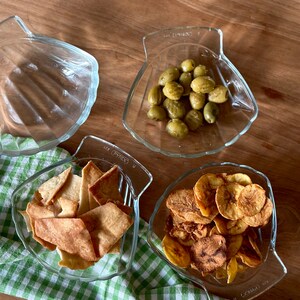 May include: Three glass bowls filled with snacks. The bowl on the left contains crispy pita chips. The middle bowl has green olives. The bowl on the right has banana chips.
