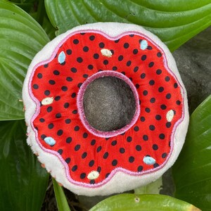May include: A red and white donut-shaped plush toy with black polka dots and a pink outline. The toy has a hole in the center.