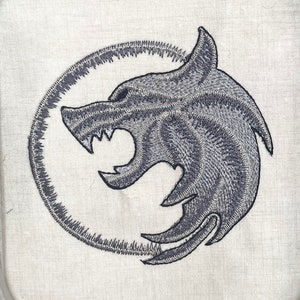 May include: Embroidered design of a gray wolf's head in a circle on a white fabric background.