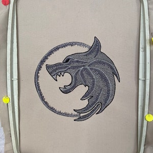 May include: Embroidered gray wolf head design in a circle on a beige fabric background.