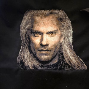May include: A close-up of a white embroidered portrait of a man with long white hair and a serious expression. His eyes are a bright yellow color.