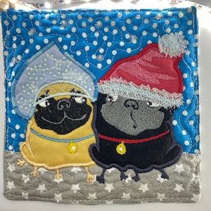 May include: Two embroidered pug dogs wearing winter hats. One pug is wearing a blue and white hat with snowflakes and the other is wearing a red and white Santa hat. The pugs are on a blue and white polka dot background with white stars.