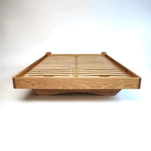 Puede incluir: Un armazón de cama de madera clara con somier de lamas. La cama tiene un diseño de perfil bajo con cabecero y piecero ligeramente elevados. El armazón está diseñado para acomodar un colchón. El armazón de la cama es de madera.