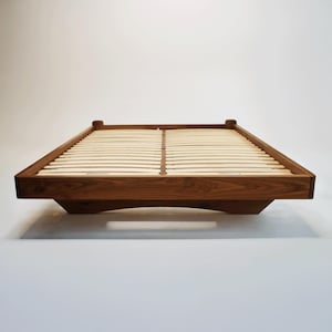 May include: A contemporary, low-profile bed frame made from dark wood. The frame has a rectangular shape with rounded corners and a slatted base. The bed frame is approximately 198 cm long and 152 cm wide.