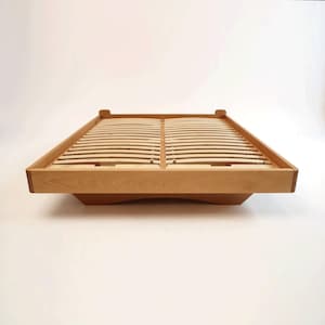 May include: A light-colored wooden bed frame with a slatted base. The frame has a low profile and a minimalist design, with a rectangular shape and rounded corners. The bed frame is made of natural wood and appears to be unfinished.