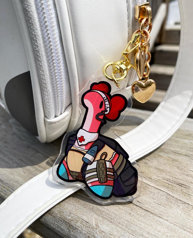 May include: A pink and red cartoon character with a medical cross on its chest and a stethoscope around its neck. The character is wearing a black vest and blue and white pants. The character is attached to a gold chain with a heart charm.