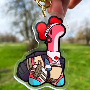 May include: A pink and red cartoon worm character wearing a white medical headband with a red cross, a black vest, and a white and blue outfit. The character is holding a medical syringe in its right hand. The character is a keychain charm made of clear acrylic.