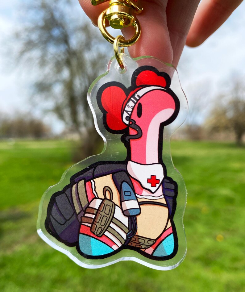 May include: A pink and red cartoon worm character wearing a white medical headband with a red cross, a black vest, and a white and blue outfit. The character is holding a medical syringe in its right hand. The character is a keychain charm made of clear acrylic.