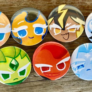 May include: A collection of seven round, colorful character button pins. Each pin features a different cartoon face with distinct features and color schemes, including blue, yellow, red, and green. The pins are arranged on a wooden surface.