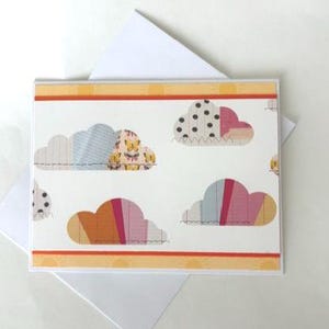 May include: A greeting card with a white background and a pattern of colorful cloud shapes. The clouds are in various colors and patterns, including polka dots and stripes. The card has an orange border and comes with a white envelope.