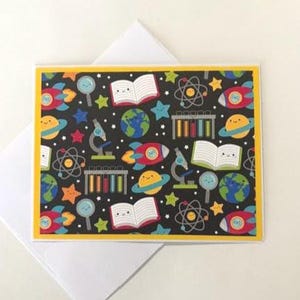 May include: Greeting card with a black background and a repeating pattern of science-themed illustrations. The design includes rockets, planets, microscopes, books, atoms, and stars. The card has a yellow border and is paired with a white envelope.