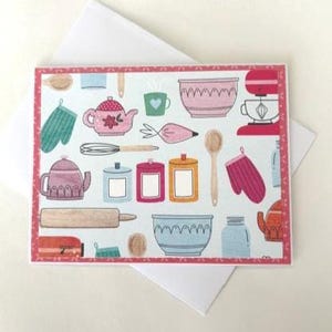 May include: A greeting card featuring a light blue background with illustrations of kitchen items. The card includes a pink teapot, a red stand mixer, a rolling pin, and a pink oven mitt. The card is on top of a white envelope.