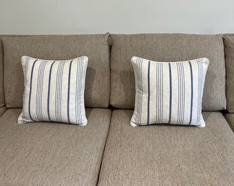 May include: Two square throw pillows with a white background and vertical navy blue stripes. The pillows have a white border and are placed on a beige sofa. The pillows add a decorative touch to the living room.