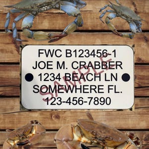 May include: A white metal tag with black text that reads "FWC B123456-1 JOE M. CRABBER 1234 BEACH LN SOMEWHERE FL. 123-456-7890". The tag is on a wooden surface with two blue crabs and two brown crabs.