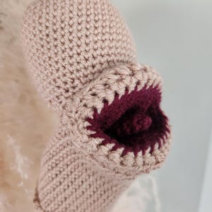 May include: A beige crocheted object with a rounded top and a protruding, open mouth-like feature. The interior of the opening is lined with a deep burgundy yarn, creating a distinct contrast against the light tan exterior of the piece.