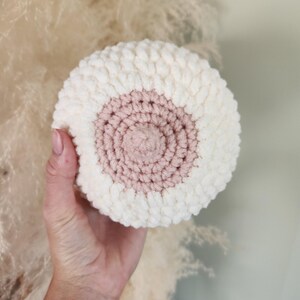 May include: A handmade, round, plush crochet item in white and light pink. The center is light pink, surrounded by a white border. The item is held in a hand against a soft, neutral background.