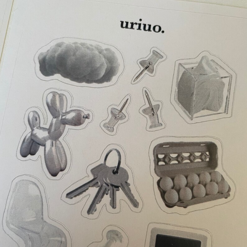 Puede incluir: Un conjunto de pegatinas grises y blancas con varios objetos, incluyendo una nube, chinchetas, un animal de globo, llaves, una caja de huevos y una caja. El texto "uriuo." est&aacute; impreso en la parte superior de la hoja.