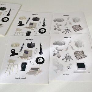 Puede incluir: Cuatro hojas de pegatinas con diferentes temas. La primera hoja tiene pegatinas en blanco y negro con una calabaza, una vela, una computadora, una silla, un clip, un cray&oacute;n y una bola de billar 8. La segunda hoja tiene pegatinas en blanco y negro con una nube, un animal de globo, un llavero, una caja de huevos y una computadora. La tercera hoja tiene pegatinas en blanco y negro con una calabaza, una vela, una computadora, una silla, un clip, un cray&oacute;n y una bola de billar 8. La cuarta hoja tiene pegatinas en blanco y negro con una nube, un animal de globo, un llavero, una caja de huevos y una computadora. El texto "uriuo." aparece en cada hoja. El texto "black mood." aparece en la primera hoja. El texto "white." aparece en la cuarta hoja.