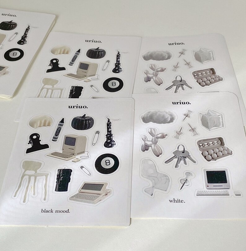 Puede incluir: Cuatro hojas de pegatinas con diferentes temas. La primera hoja tiene pegatinas en blanco y negro con una calabaza, una vela, una computadora, una silla, un clip, un cray&oacute;n y una bola de billar 8. La segunda hoja tiene pegatinas en blanco y negro con una nube, un animal de globo, un llavero, una caja de huevos y una computadora. La tercera hoja tiene pegatinas en blanco y negro con una calabaza, una vela, una computadora, una silla, un clip, un cray&oacute;n y una bola de billar 8. La cuarta hoja tiene pegatinas en blanco y negro con una nube, un animal de globo, un llavero, una caja de huevos y una computadora. El texto "uriuo." aparece en cada hoja. El texto "black mood." aparece en la primera hoja. El texto "white." aparece en la cuarta hoja.