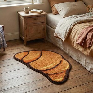 May include: A brown and orange rug shaped like a croissant rests on a wooden floor. The rug has a dark brown border and lighter orange and yellow details. The rug is in a bedroom setting.