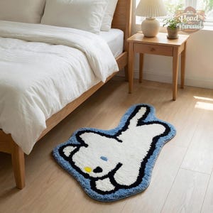 May include: A white and blue cartoon bunny rug with a black outline. The rug is on a light wood floor next to a bed with white bedding and a wooden nightstand with a lamp.