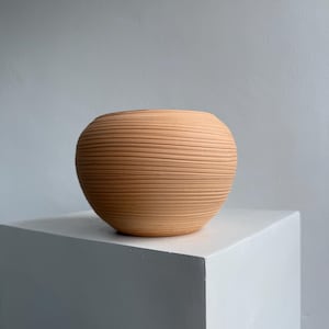 May include: A terracotta-colored, spherical vase with horizontal, ridged texture. The vase sits on a white, square pedestal against a gray background. The rounded shape and textured surface create a modern, minimalist aesthetic, suitable for home decor.