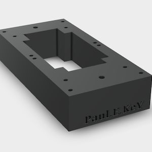 Op de afbeelding: Een donkergrijze rechthoekige blok met een verzonken midden en meerdere geboorde gaten. De blok heeft de gegraveerde tekst "Paul E. Key". Het ontwerp suggereert een structurele of mechanische component.