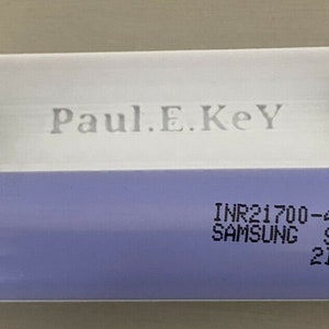 Może przedstawiać: Biały plastikowy uchwyt na baterie z srebrnym metalowym zatrzaskiem po obu stronach. Uchwyt zawiera fioletową cylindryczną baterię z nadrukiem "INR21700-40T SAMSUNG SDI 21A4".