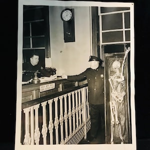 May include: Black and white photograph of a police officer standing next to a skeleton in a glass case. Another officer is seated behind a counter. A clock hangs on the wall. The railing has the text "KEEP OFF RAIL STAND OUTSIDE RAIL".
