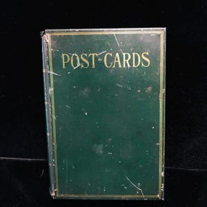 May include: A vintage green metal postcard holder with the words "POST CARDS" in gold lettering. The holder has a rectangular shape and shows signs of wear, with scratches and minor imperfections.