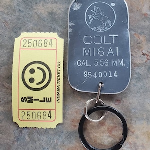 May include: A silver metal keychain with a black ring. The keychain has a Colt M16A1 logo with the text "CAL. 5.56 MM." and the serial number "9540014".