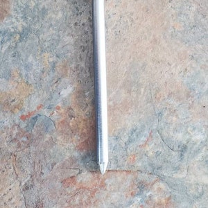 Pode incluir: Uma haste de metal prateada com uma ponta arredondada e uma parte superior com cristas. A haste mede aproximadamente 15 cm de comprimento.