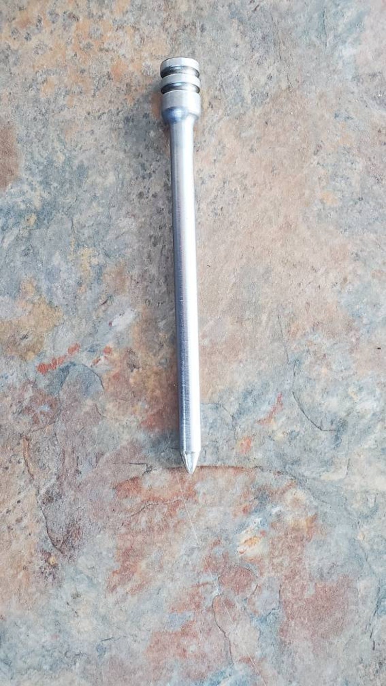 Pode incluir: Uma haste de metal prateada com uma ponta arredondada e uma parte superior com cristas. A haste mede aproximadamente 15 cm de comprimento.