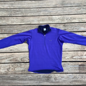 May include: A purple long-sleeved shirt with a half-zip closure and a small logo on the chest.