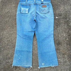 May include: A pair of light blue denim jeans with a wide leg opening. The jeans have a brown leather patch on the back with the word "Wrangler" printed on it. The jeans have a frayed hem.