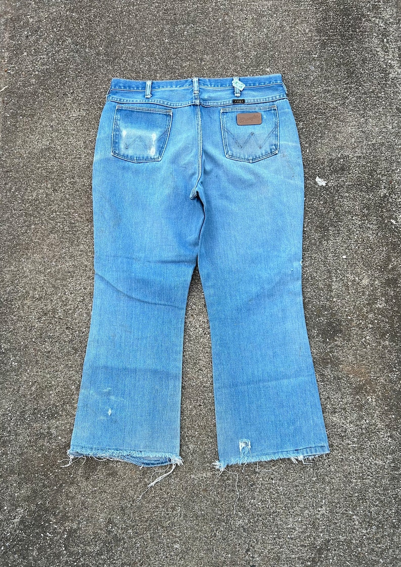 May include: A pair of light blue denim jeans with a wide leg opening. The jeans have a brown leather patch on the back with the word "Wrangler" printed on it. The jeans have a frayed hem.