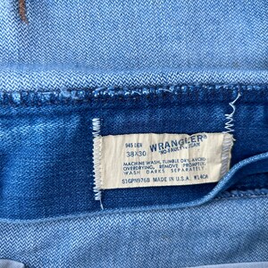 May include: A blue denim label with white text that reads "945 DEN 38X30 WRANGLER&reg; NO-FAULT JEAN MACHINE WASH, TUMBLE DRY, AVOID OVERDRYING, REMOVE PROMPTLY WASH DARKS SEPARATELY S1GPN976B MADE IN U.S.A. W14CA"