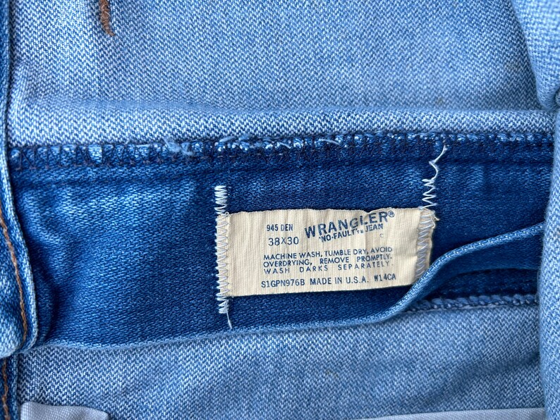 May include: A blue denim label with white text that reads "945 DEN 38X30 WRANGLER&reg; NO-FAULT JEAN MACHINE WASH, TUMBLE DRY, AVOID OVERDRYING, REMOVE PROMPTLY WASH DARKS SEPARATELY S1GPN976B MADE IN U.S.A. W14CA"