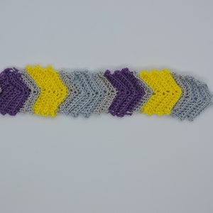 May include: A handmade crochet bracelet in a chevron pattern. The bracelet features alternating sections of purple, yellow, and gray yarn. A silver-colored button is attached to the purple section. The bracelet has a loop at the end.