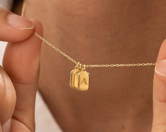 May include: A gold-toned necklace featuring two small, rectangular pendant charms suspended from a delicate chain. One of the charms is engraved with the capital letter A, while the other charm remains plain and smooth in a polished metallic finish.