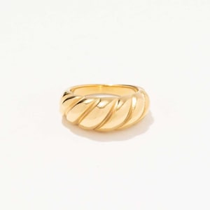 May include: A gold-colored ring with a thick, rounded band. The ring features a ridged design that wraps around the entire band. The ring is set against a white background, highlighting its shiny, reflective surface. Jewelry.